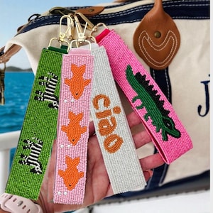 May include: A collection of beaded keychains in various colors. One green keychain features a zebra design, a pink one has orange fish, a white one says "ciao", and a pink one has a green alligator.