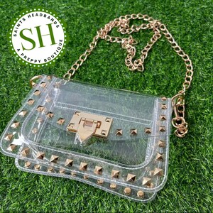 May include: Clear crossbody bag with gold-colored chain strap and studded accents. The bag features a gold-colored clasp and is set against a green grass background. The bag is from Saints Headbands A Preppy Boutique.