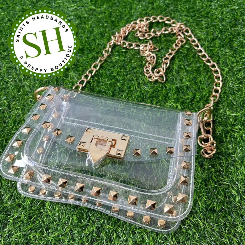 May include: Clear crossbody bag with gold-colored chain strap and studded accents. The bag features a gold-colored clasp and is set against a green grass background. The bag is from Saints Headbands A Preppy Boutique.