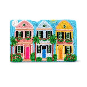 May include: A rectangular beaded clutch featuring a vibrant scene of three colorful houses in pink, white, and yellow. The design includes green palm trees, red flower boxes, a bright blue sky with a yellow sun, and a white moon against a blue background.