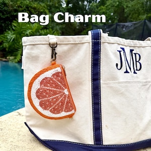 K&ouml;nnte beinhalten: Eine Canvas-Tragetasche mit marineblauem Besatz und dem gestickten Monogramm JMB, an deren Griff ein perlenbesetzter Taschenanh&auml;nger in Form einer Orangenscheibe mit einem Metallverschluss befestigt ist. Der Anh&auml;nger zeigt ein Zitrussegment.