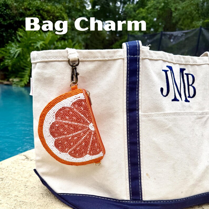 K&ouml;nnte beinhalten: Eine Canvas-Tragetasche mit marineblauem Besatz und dem gestickten Monogramm JMB, an deren Griff ein perlenbesetzter Taschenanh&auml;nger in Form einer Orangenscheibe mit einem Metallverschluss befestigt ist. Der Anh&auml;nger zeigt ein Zitrussegment.