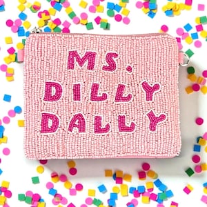 May include: A pink beaded clutch with the words "MS. DILLY DALLY" in bright pink letters. The clutch has a zipper closure and a small loop on the side. Colorful confetti in various shapes and sizes surrounds the clutch.