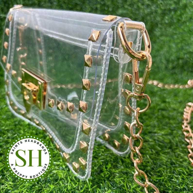 May include: A clear, rectangular crossbody bag with gold-colored hardware and studded accents. The bag features a gold chain strap and a rectangular clasp. The logo for "Saints Headbands A Preppy Boutique" is visible.