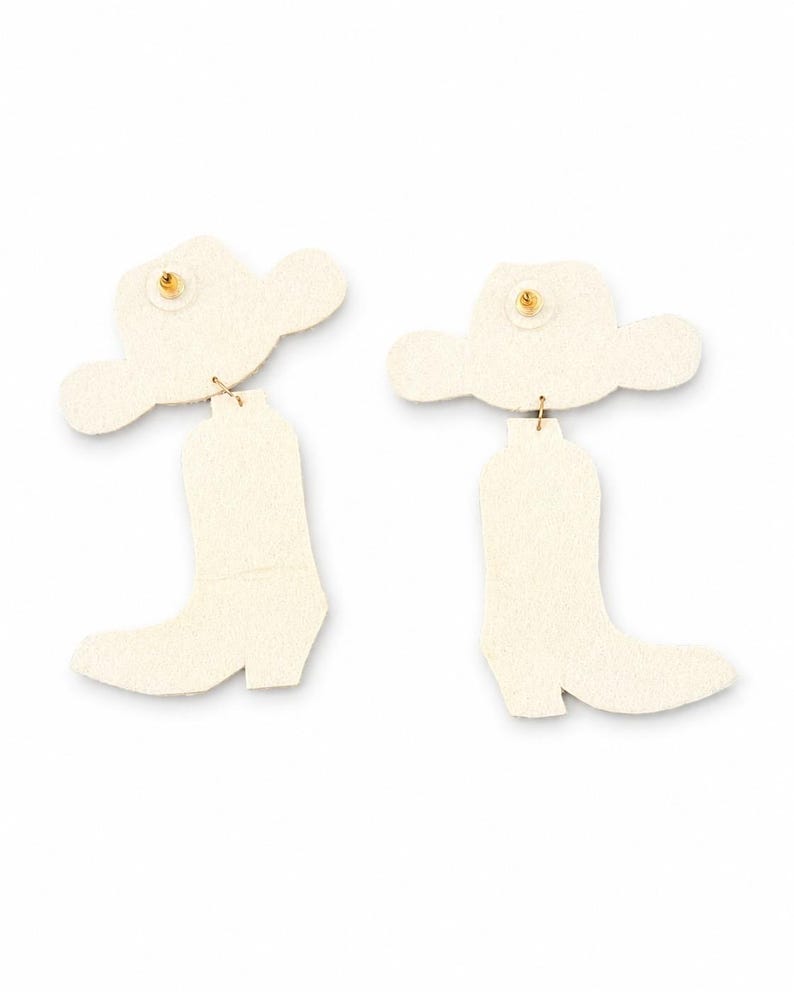 May include: A pair of off-white earrings featuring a cowboy hat top piece connected by a small metal jump ring to a cowboy boot bottom piece. The back of each earring shows a gold-toned metal post backing secured to the center of the hat.