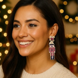 Op de afbeelding: Een vrouw draagt feestelijke, bungelende oorbellen. De oorbellen hebben de vorm van een notenkrakersoldaat, met rode, gouden en zwarte accenten. De achtergrond is wazig met kerstverlichting, wat wijst op een vakantiethema.