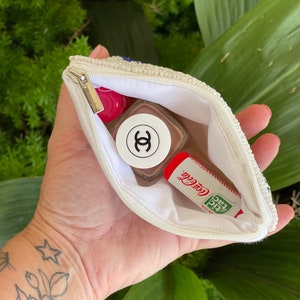 May include: A white beaded zippered pouch with a pink and white nail polish bottle and a red and green lip balm inside. The lip balm has the text "Coca-Cola" and "Edition Special" printed on it.