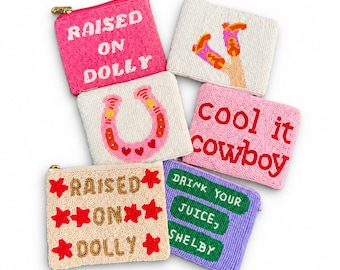 Peut inclure: Six pochettes zippées rectangulaires en perles avec divers motifs : une pochette rose avec "RAISED ON DOLLY", une blanche avec des bottes de cow-boy, une blanche avec un fer à cheval, une rose avec "cool it cowboy", une beige avec "RAISED ON DOLLY" et des étoiles rouges, et une violette avec "DRINK YOUR JUICE, SHELBY".