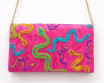 May include: A rectangular hot pink clutch bag featuring a sequined surface and a pattern of beaded snakes in vibrant shades of yellow, lime green, purple, and turquoise. The bag includes a thin gold-toned metal chain strap attached at the top corners.
