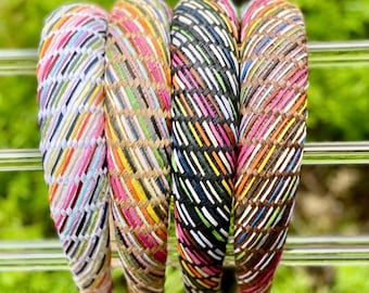 May include: Four colorful woven headbands. Each headband features a unique pattern of diagonal stripes in various colors, including pink, orange, green, and white. The headbands are displayed against a blurred green background.