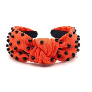 May include: Orange knotted headband with black beads. The headband is made of fabric and has a knotted design. The beads are evenly spaced around the headband.