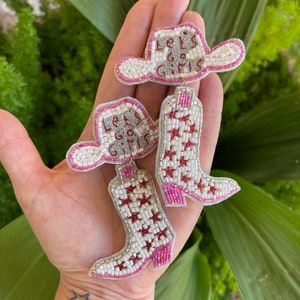 May include: A pair of beaded earrings featuring a white cowboy hat with the text "Let's Go Girls" in pink and silver beads, attached to a white cowboy boot with pink and silver star patterns and a pink heel, crafted with intricate beadwork.