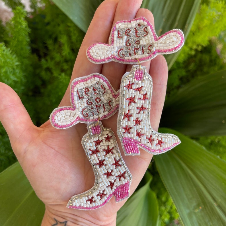 May include: A pair of beaded earrings featuring a white cowboy hat with the text "Let's Go Girls" in pink and silver beads, attached to a white cowboy boot with pink and silver star patterns and a pink heel, crafted with intricate beadwork.