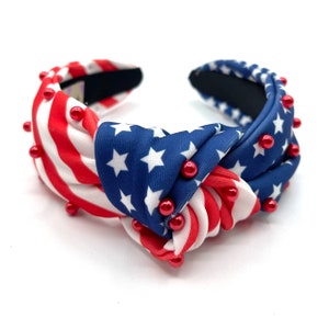 May include: A patriotic headband featuring a knotted design. The headband is adorned with red, white, and blue fabric, mimicking the American flag. Small red beads are evenly spaced along the sides, adding a decorative touch.