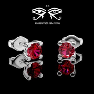 May include: A pair of silver stud earrings featuring round-cut, vibrant red gemstones held by four-prong settings. Above the earrings, the text I.C. IMAGINEERED CREATIONS appears alongside a stylized graphic of two eyes with decorative lines.