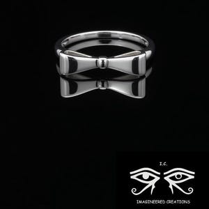 May include: A polished silver ring featuring a bow-tie design. The ring is reflected on a black surface. The image also includes a logo with stylized eyes and the text "IMAGINEERED CREATIONS".