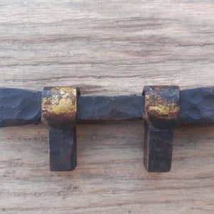 May include: A hand-forged metal cabinet pull featuring a dark, hammered texture with two prominent gold-toned accents wrapping around the horizontal bar. The hardware includes two vertical mounting posts extending downward from the main handle.