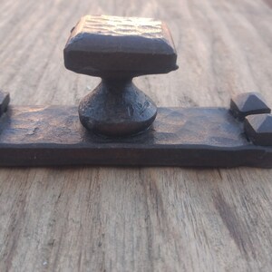 May include: A dark brown, hand-forged metal cabinet pull featuring a central square knob with a hammered texture. The knob is mounted on a rectangular base plate that includes two small, pyramid-shaped decorative studs on each end of the plate.