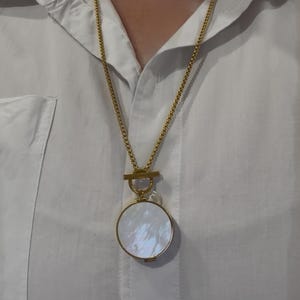 May include: A gold-toned necklace with a round, iridescent pendant. The pendant is suspended from a delicate chain and features a mother-of-pearl effect. The necklace is worn over a white button-down shirt.