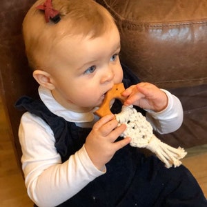 May include: A baby wearing a white long-sleeved shirt and a dark blue overall is holding a wooden teether with a white crocheted string attached. The baby has a red bow clip in their hair.