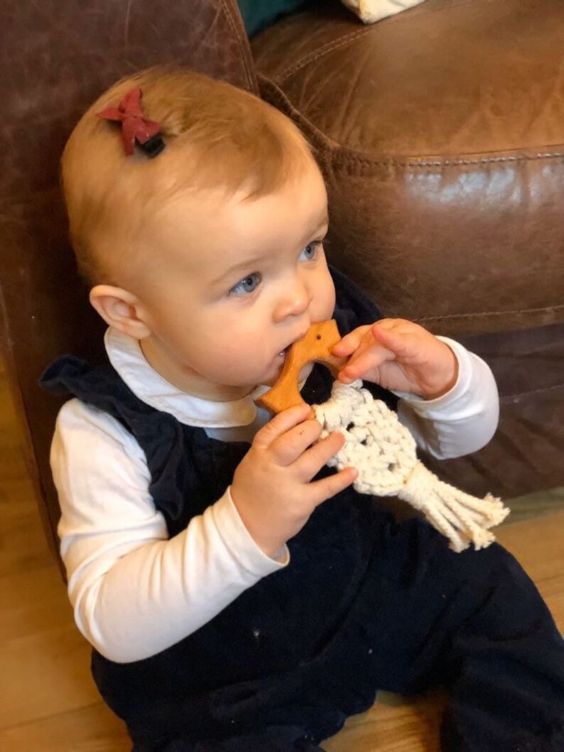May include: A baby wearing a white long-sleeved shirt and a dark blue overall is holding a wooden teether with a white crocheted string attached. The baby has a red bow clip in their hair.