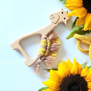 Puede incluir: Un mordedor de madera con forma de jirafa con un borla de macramé amarillo y beige. El mordedor está rodeado de girasoles.