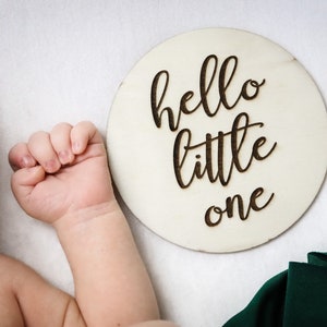 以下が含まれることがあります： 黒い筆記体で「hello little one」と書かれた白い木製の円形