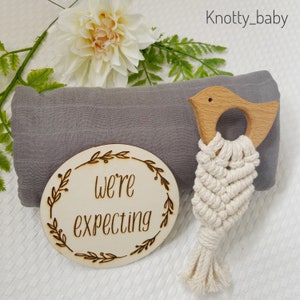 May include: A gray muslin swaddle blanket with a wooden bird teether and a wooden circle with the text "We're expecting" on it. The circle is decorated with a floral design.