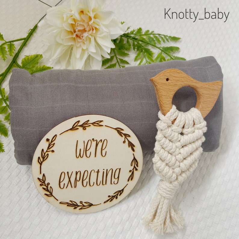 May include: A gray muslin swaddle blanket with a wooden bird teether and a wooden circle with the text "We're expecting" on it. The circle is decorated with a floral design.