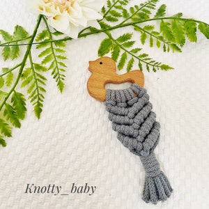 Op de afbeelding: Een grijs macramé babyspeelgoed met een houten eendenkop eraan bevestigd. Het speelgoed heeft een lange kwast aan de onderkant.