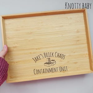 Könnte beinhalten: Ein rechteckiges Tablett aus Bambus mit erhöhten Kanten. Das Tablett trägt den Text "JAKE'S BRICK CHAOS CONTAINMENT UNIT" und eine Grafik von Spielzeugblöcken. Die Worte "KNOTTY BABY" sind oben rechts aufgedruckt.