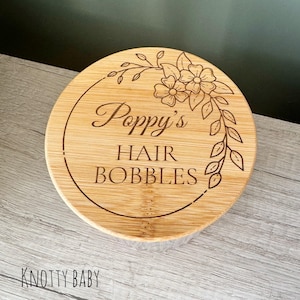 May include: A round wooden container with a floral design and the text "Poppy's Hair Bobbles" engraved on the lid.