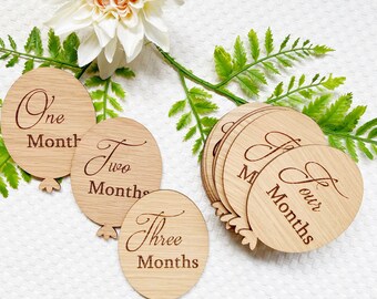 Può includere: Carte di legno per i traguardi del bambino a forma di palloncini con le parole "Un mese", "Due mesi", "Tre mesi" e "Quattro mesi" incise su di esse. Le carte sono disposte su una superficie bianca con una pianta verde sullo sfondo. Il testo "Knotty Baby" è visibile nell'angolo in basso a destra.