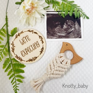 May include: A wooden circle with the text "We're expecting" surrounded by a floral design. A black and white ultrasound image is in the background. A wooden bird-shaped teether with a white macrame design is in the foreground.