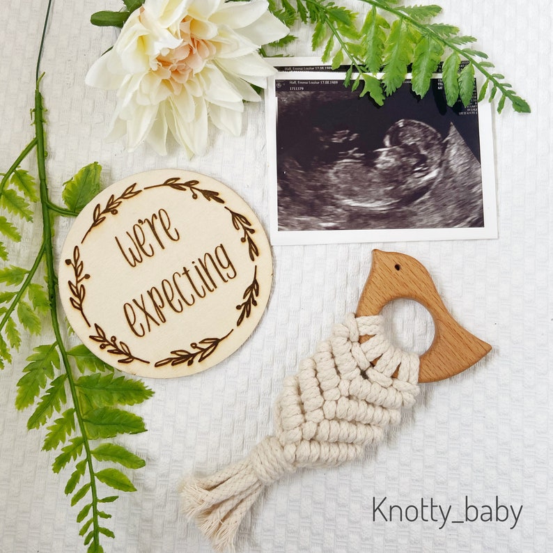 May include: A wooden circle with the text "We're expecting" surrounded by a floral design. A black and white ultrasound image is in the background. A wooden bird-shaped teether with a white macrame design is in the foreground.