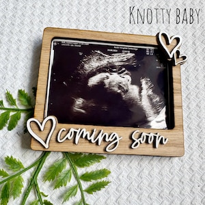 Puede incluir: Un marco de fotos de madera con fondo blanco y el texto "Knotty Baby coming soon". El marco tiene un recorte de dos corazones y una imagen de sonograma en el interior.