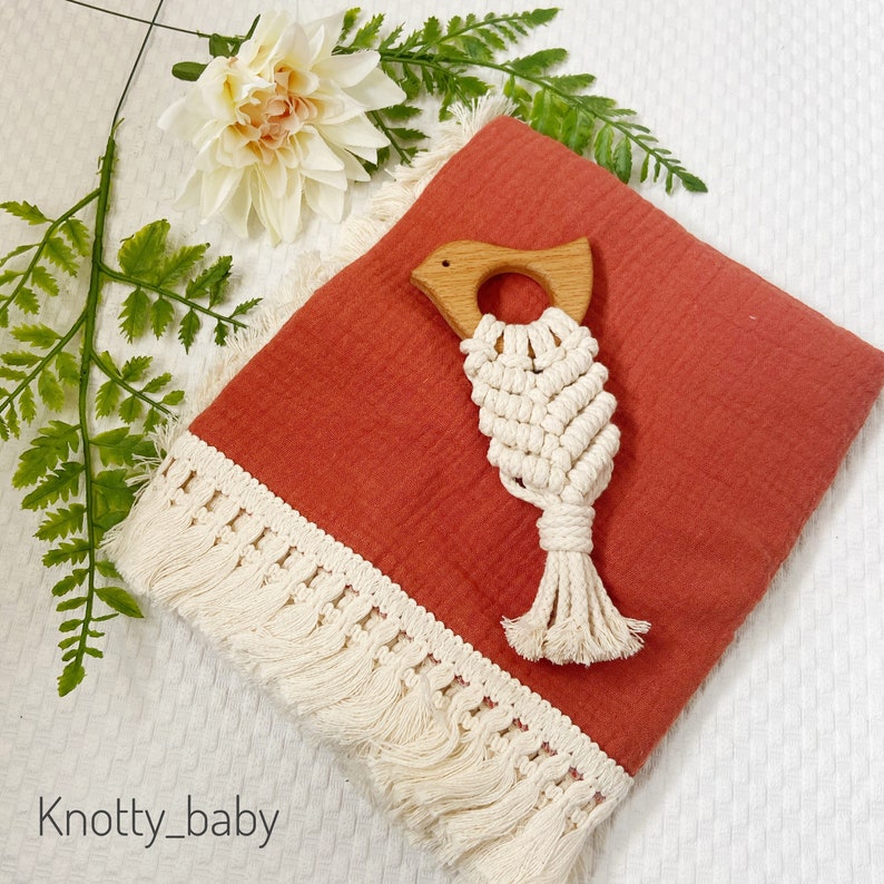 May include: A terracotta muslin baby blanket with a white fringe border and a wooden bird teether with a macrame tassel.