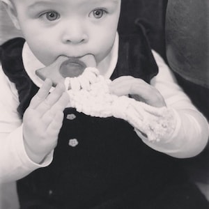 May include: A baby in a black velvet dress is holding a wooden teether with a crocheted strap. The teether is shaped like a triangle with rounded edges.