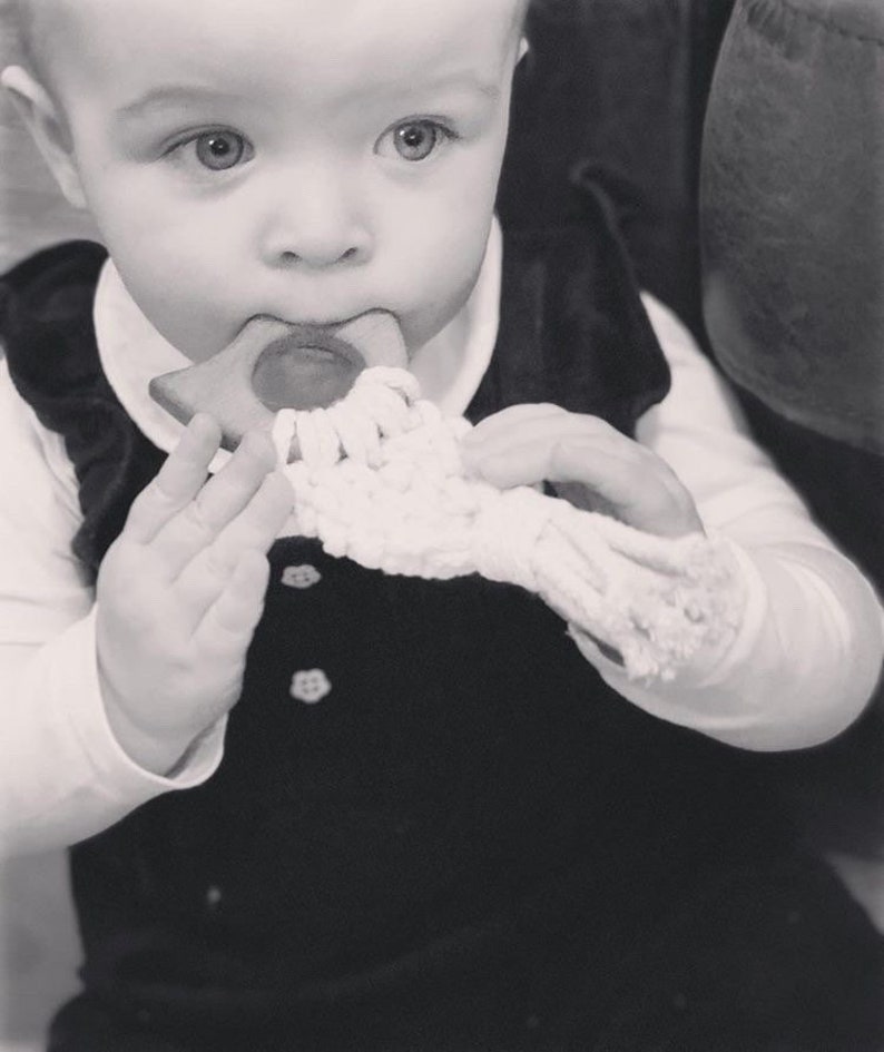 May include: A baby in a black velvet dress is holding a wooden teether with a crocheted strap. The teether is shaped like a triangle with rounded edges.
