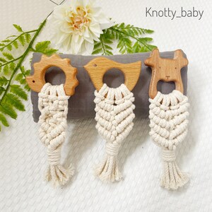 May include: Three wooden teethers with knotted macrame tassels. The teethers are shaped like a bird, a hedgehog, and a dog. The tassels are a cream color and the teethers are a natural wood color.