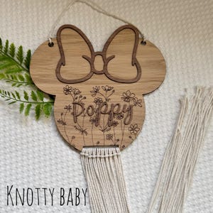 May include: Wooden cutout of a mouse head with a bow and floral design. The name "Poppy" is engraved on the cutout. A white macrame tassel hangs from the bottom.