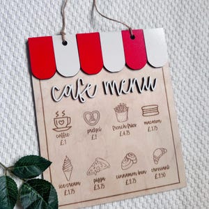May include: Wooden cafe menu sign with a red and white awning design. The menu includes coffee, pretzels, fries, macarons, ice cream, pizza, cinnamon buns, and croissants. The sign is made of light-colored wood and has a string for hanging.