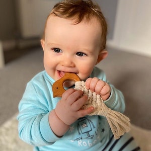 May include: A baby in a blue and white striped onesie is holding a wooden teether shaped like a bird. The teether has a beige rope attached to it.