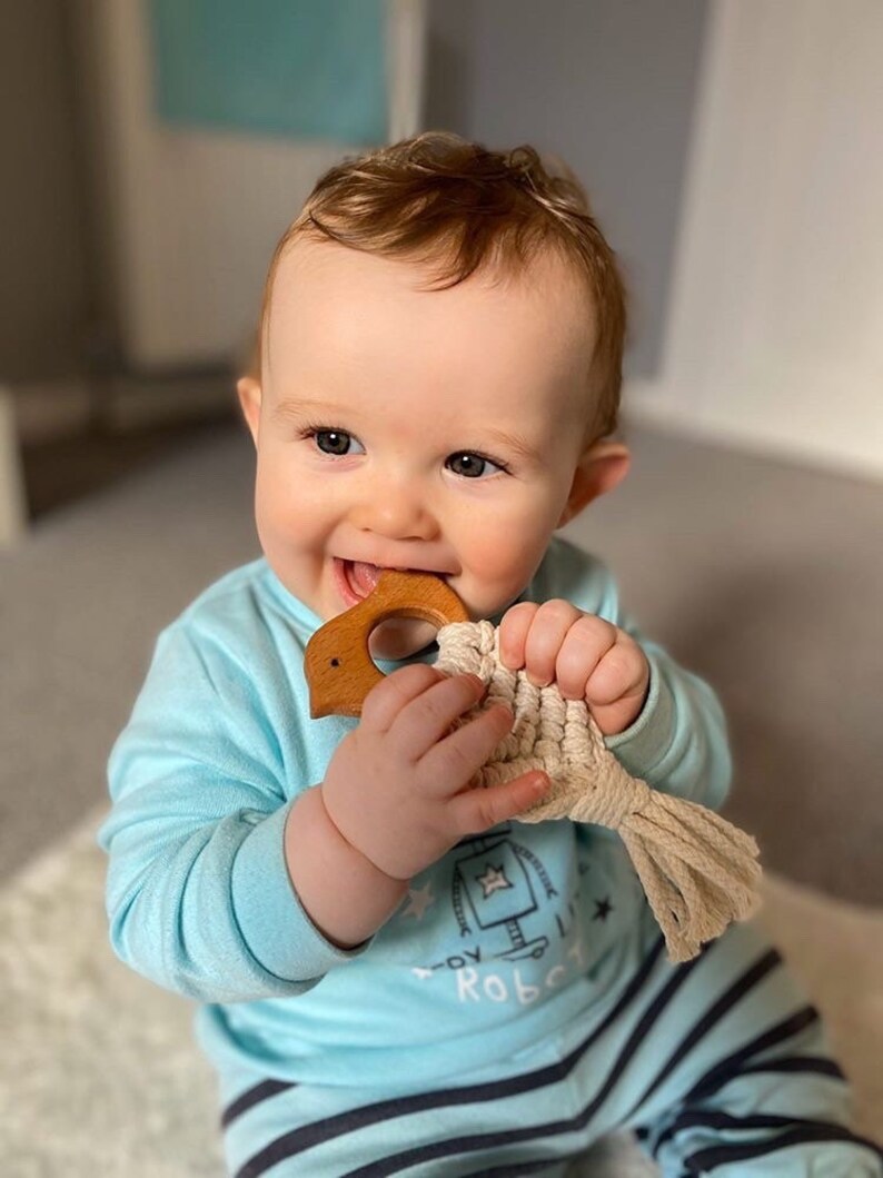May include: A baby in a blue and white striped onesie is holding a wooden teether shaped like a bird. The teether has a beige rope attached to it.