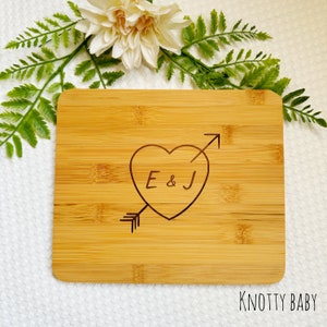 May include: A natural bamboo cutting board with a heart design and arrow engraved with the initials "E & J". The board is a light brown color and has a smooth surface. The text "Knotty Baby" is printed in the bottom right corner.