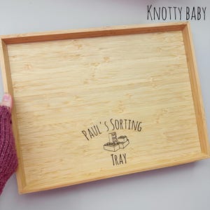 May include: A rectangular bamboo tray with a raised edge. The tray has the text "PAUL'S SORTING TRAY" and a graphic of toy blocks. The text "KNOTTY BABY" is in the upper right corner.