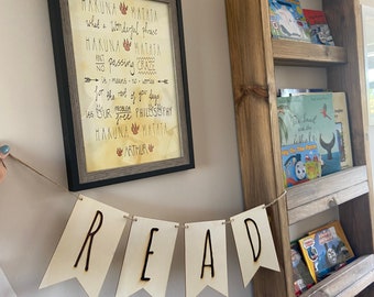 Könnte beinhalten: Ein Holzschild mit dem Spruch "Hakuna Matata" und einem Zitat über die Bedeutung des Spruchs. Das Schild hängt an einer Wand mit einem Holzregal daneben. Ein Holzbanner mit dem Wort "READ" hängt unter dem Schild.