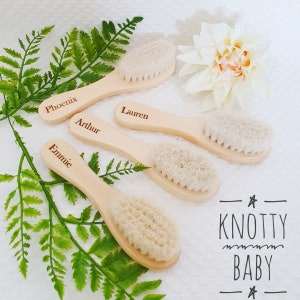 May include: Four wooden baby hair brushes with natural bristles. Each brush has a name engraved on the handle: Phoenix, Arthur, Lauren, and Emmie. The brushes are arranged on a white surface with green ferns and a white flower.