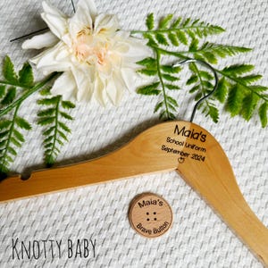 Puede incluir: Una percha de madera con el texto "Maia's School Uniform September 2024" grabado en ella. Un botón de madera con el texto "Maia's Brave Button" está unido a la percha.
