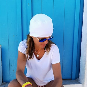 Könnte beinhalten: Eine Frau in einem weißen T-Shirt, einer weißen Mütze und einer blauen Sonnenbrille sitzt auf einer weißen Stufe vor einer blauen Tür. Sie trägt einen bunten Bikini-Unterteil und ein gelbes Armband.